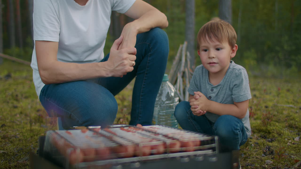 En voksen mand og en lille dreng sidder ved en kulgrill med kød og pølser under en familietur i skoven. Far og søn.