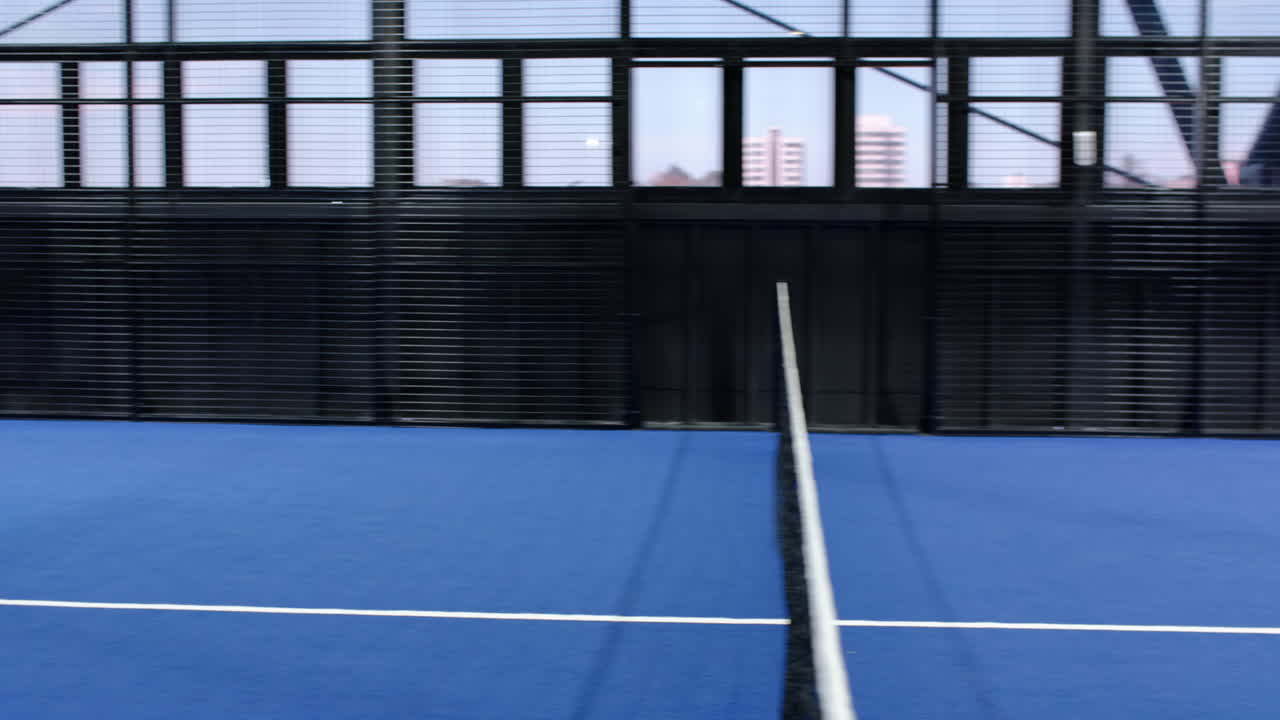 Empty padel indoor court with blue flooring and cityscape view through large windows