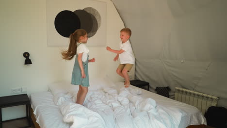 children have fun on double bed at glamping. playful little boy with elder sister hop on mattress in bedroom. happy kids jump in camping tent