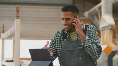 Male-Apprentice-With-Digital-Tablet-Working-As-Carpenter-In-Furniture-Workshop-Talking-On-Mobile-Phone