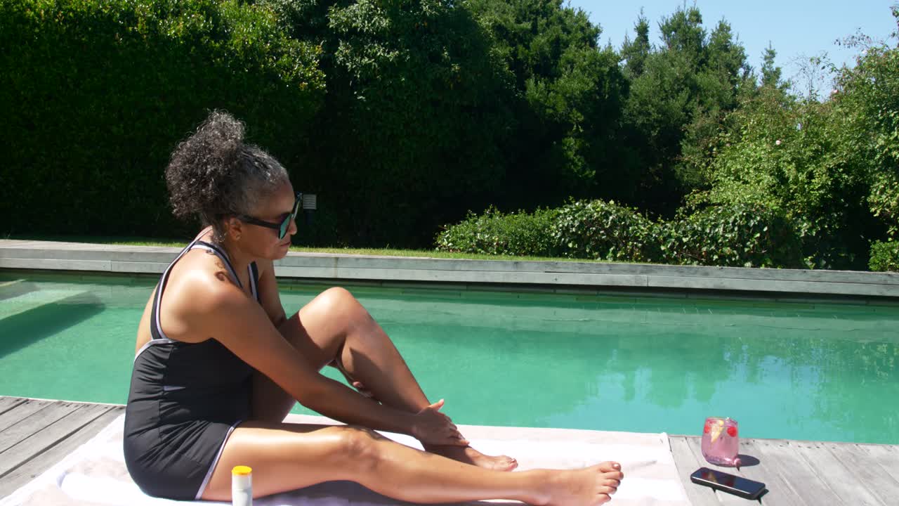 Eine afroamerikanische Frau greift nach Sonnencreme und reibt sich am Pool die Beine als Sonnenschutz ein, mit Platz für Text.