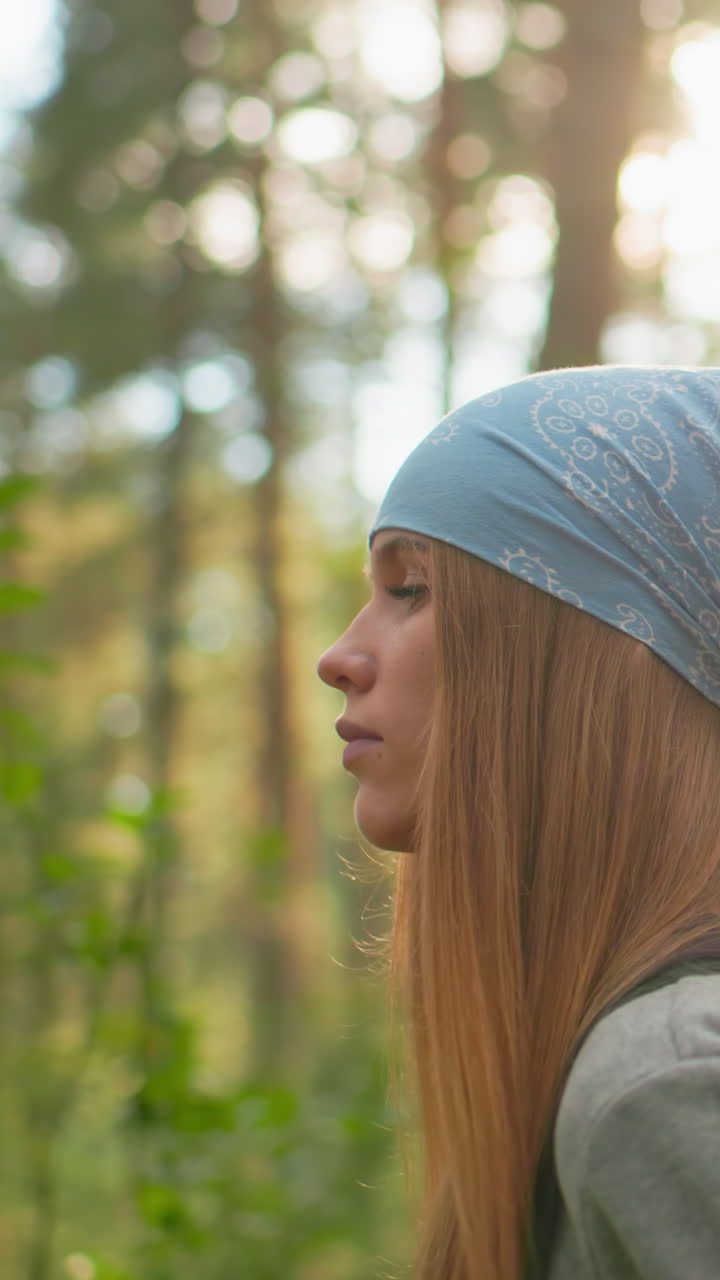 dama con pañuelo azul y sudadera gris camina a través del bosque iluminado por el sol, camina pensativa, su compañera, con camisa verde, la sigue de cerca, rodeada de vegetación y suave luz solar