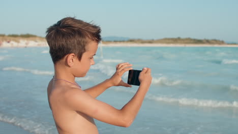 niño alegre buscando una pantalla en la playa. adolescente filmando en la cámara en la playa.