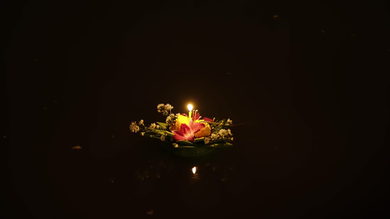 A delicate floating lantern with flowers and candle drifts across the dark river during Yi Peng in Chiang Mai, Thailand.