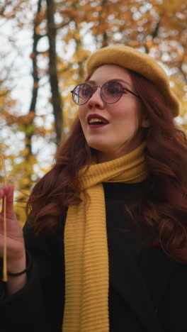 elegante dama con boina amarilla girando hoja de otoño en la mano mientras pasea por un parque vibrante rodeado de follaje dorado, disfrutando de la naturaleza pacífica y el aire fresco de otoño con una sonrisa graciosa