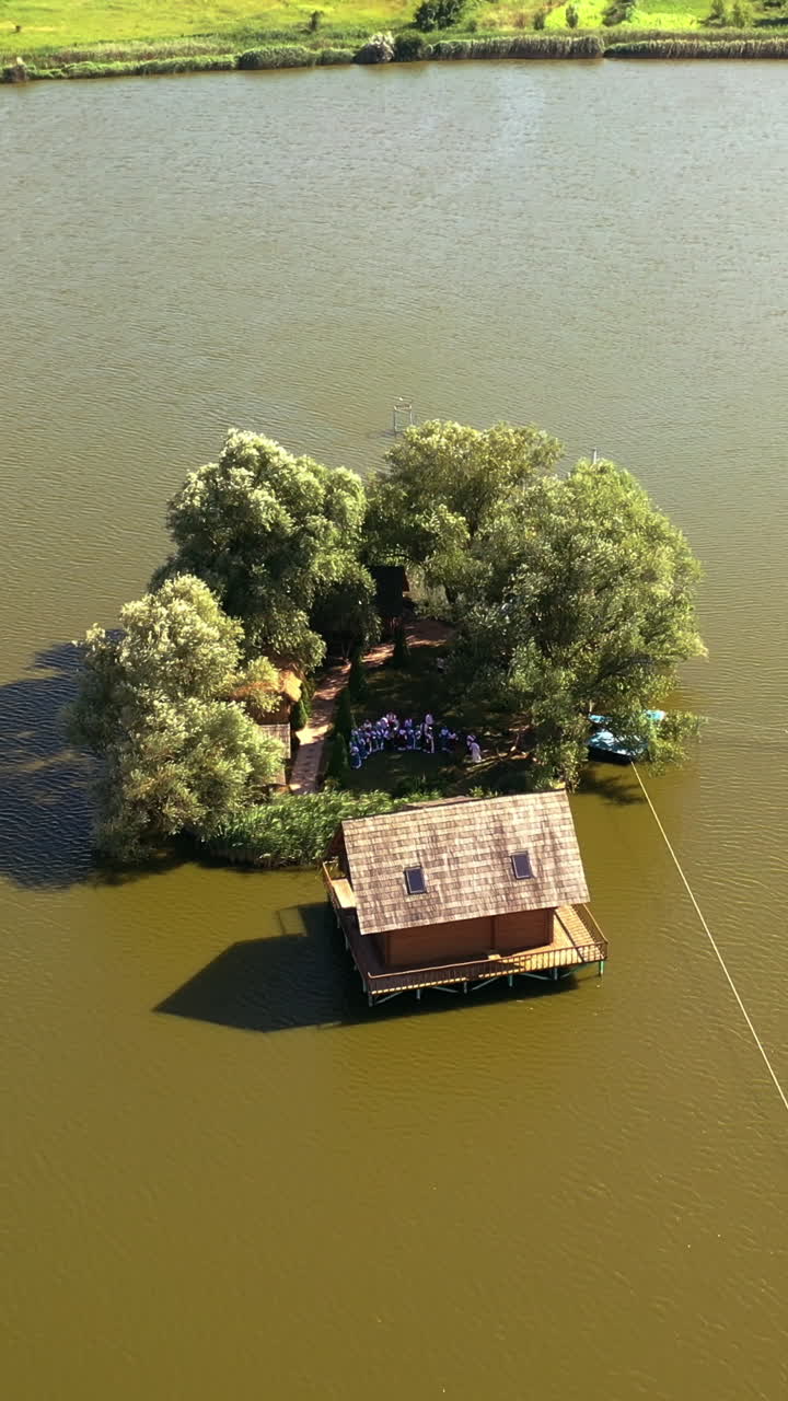 Small island with green trees and a house in the river. Aerial view of island and wooden house in the middle of a lake surrounded by water at daytime in summer. Camera moves down. Vertical video