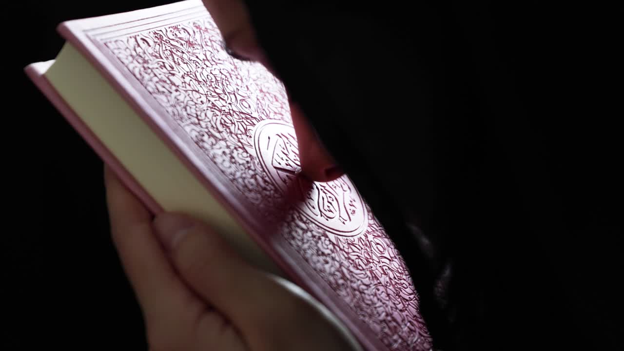 A Muslim woman in hijab leans her forehead to a pink Quran, she raises her head and kisses the Quran out of respect and attachment to it