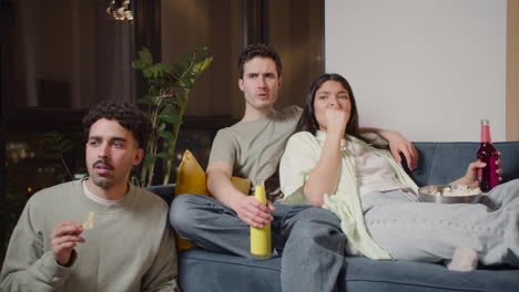 couple and a friend watching unpleasant movie scene on television sitting on couch and floor, eating popcorn and drinking soda