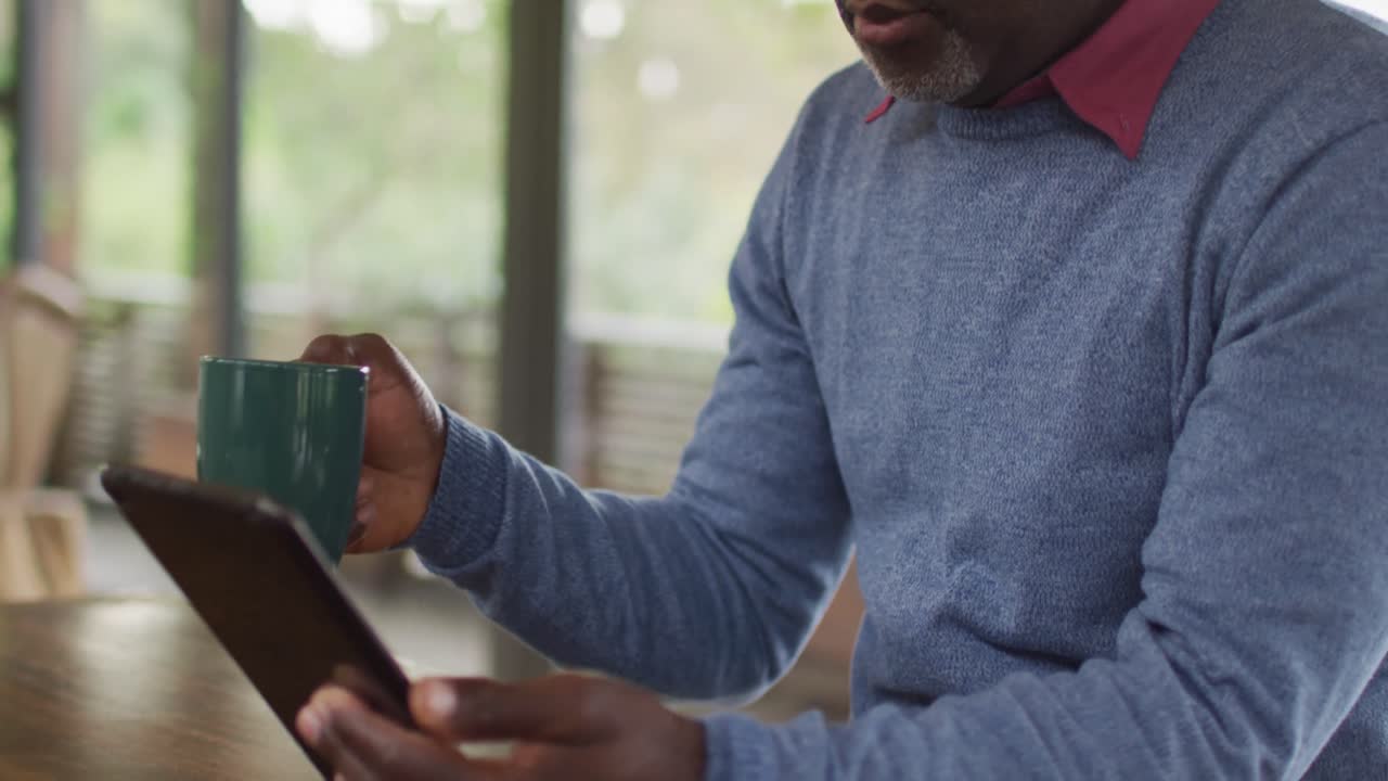 African american senior man sitting at counter in kitchen using tablet and drinking coffee