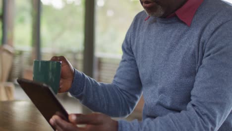 African-american-senior-man-sitting-at-counter-in-kitchen-using-tablet-and-drinking-coffee