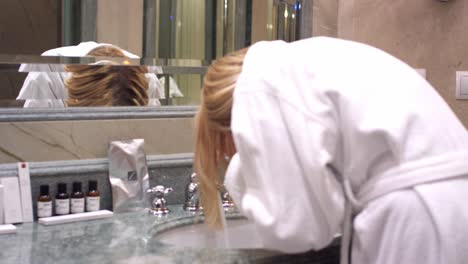 woman in white bathrobe washing face in bathroom sink room back view