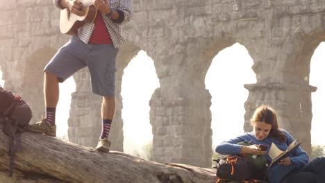 happy young couple backpackers tourists on a log trunk playing guitar singing reading book in front of ancient roman aqueduct ruins in romantic parco degli acquedotti park in rome at sunrise slow motion