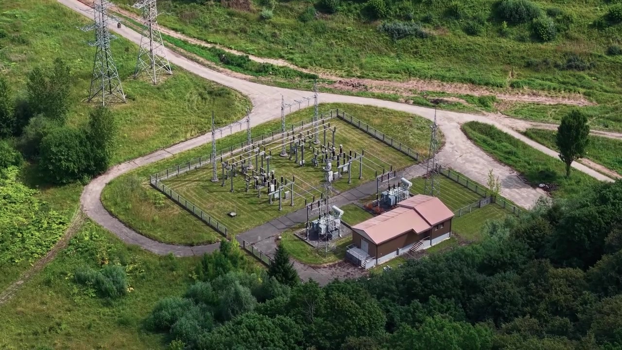 Video z dronu odhaluje širokoúhlý letecký záběr venkovní elektrické rozvodny, elektrického vedení, přístupových cest a svěží zeleně za přirozeného denního světla. Venkovní, průmyslová, statická scéna