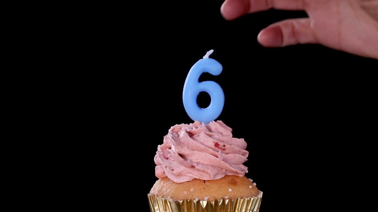 A hand inserts a blue number six birthday candle into a strawberry-frosted cupcake against a black background, with steady lighting and minimal camera movement