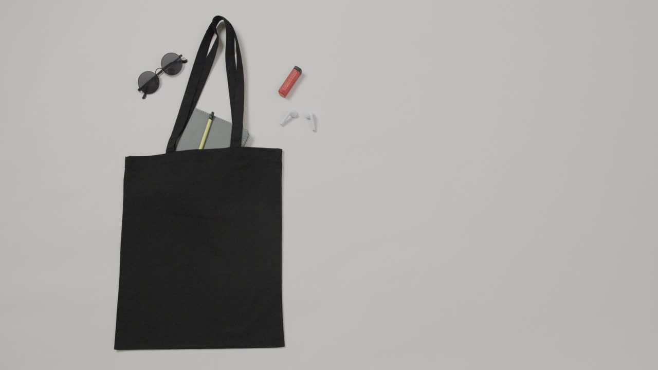 Borsa nera con occhiali da sole, taccuino, rossetto su sfondo bianco, spazio per il testo, al rallentatore
