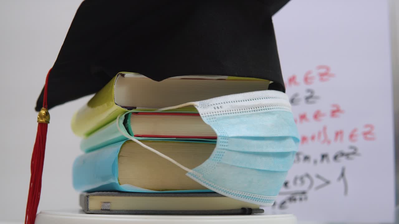 보호 의료 마스크와 대학 졸업생의 검은 사각형 모자가 기둥으로 접힌 책에 붙어 있습니다.