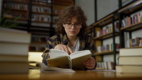 confident girl student with curly hair wearing glasses reads a book among books at a table at the university