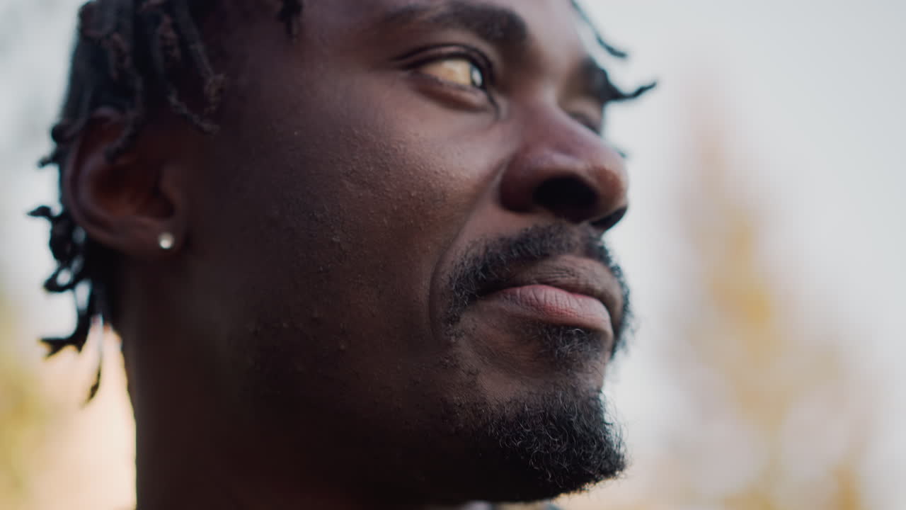 Primer plano de un hombre negro comiendo un sándwich en un parque al aire libre durante la hora dorada. Los primeros planos cinematográficos íntimos muestran la piel texturizada, las rastas, los pendientes, las migas y el envoltorio de papel, con una mirada lenta y pensativa.