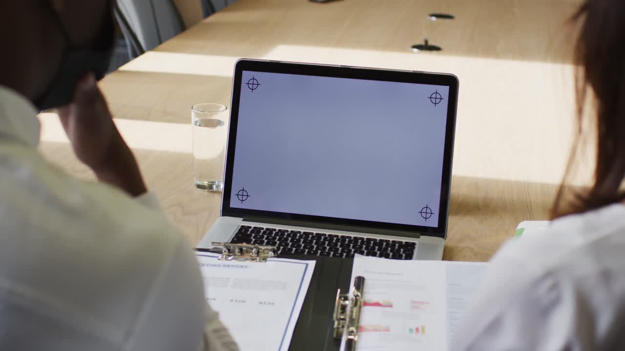 Diverse businessman and businesswoman wearing face masks using laptop with blank screen