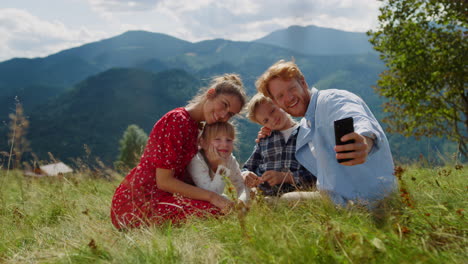 positive family taking selfie on cellphone sitting green grass mountain hill.