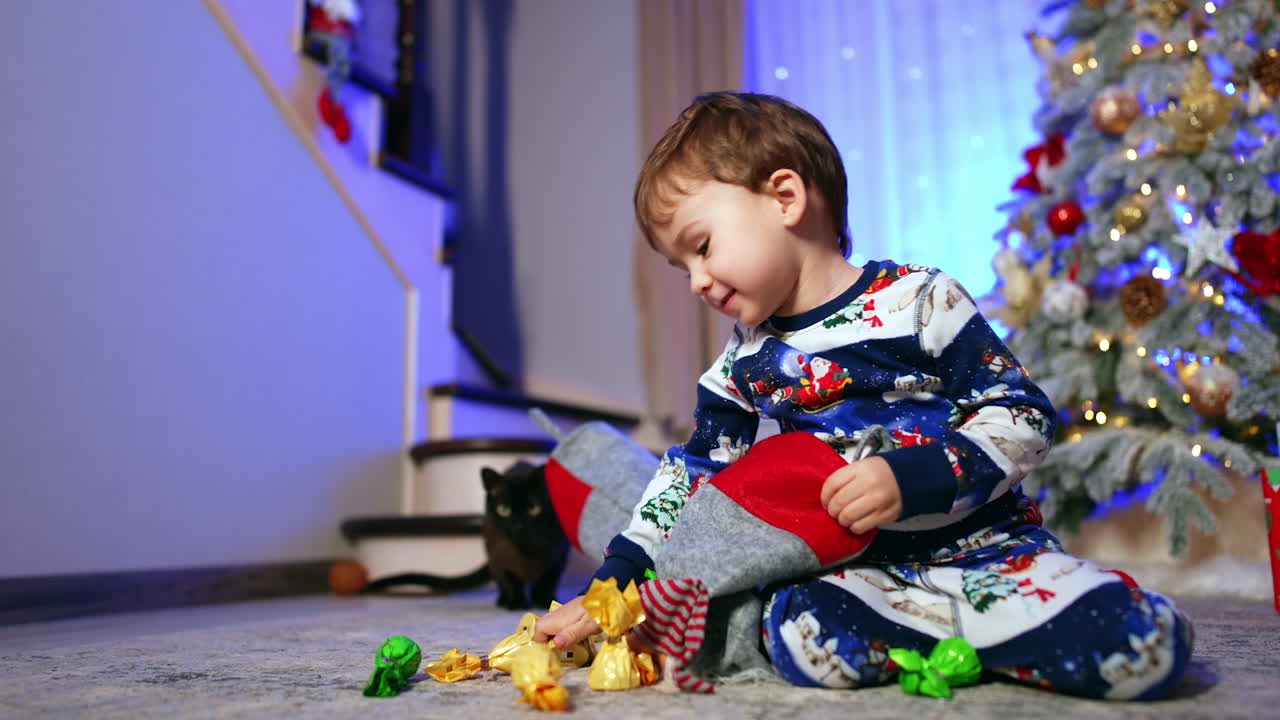 Small cute kid plays with Christmas stocking near the Christmas tree. Happy baby gets candies and black cat watches him.