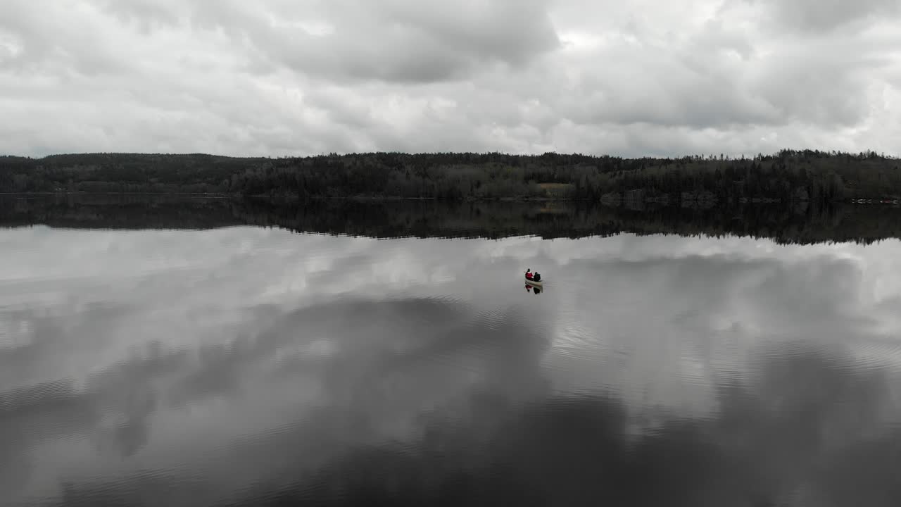 dolly en toma aérea descendente de dos personas remando suavemente en una canoa blanca mientras crea pequeñas ondas en todo un tranquilo lago reflectante hacia tierra en un día nublado gris