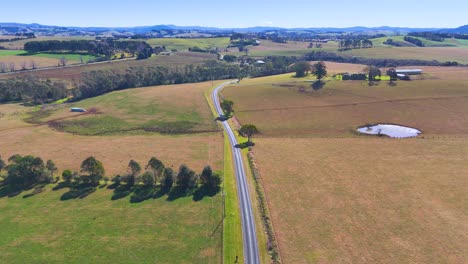 drone footage glides above a quiet country road, passing open fields, scattered trees, and small ponds under bright daylight in rural new south wales