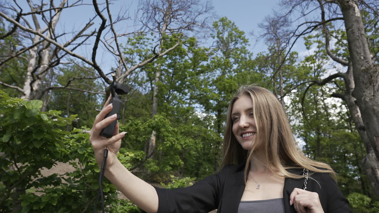 화창한 여름날 녹색 숲에서 셀카 비디오를 만드는 카메라가 있는 작은 짐벌을 가진 젊은 여성, 여행하는 동영상 블로거 개념, 느린 동작