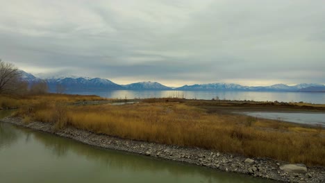 flying low over a field and river that empties into a large reflective lake with mountains in the distance