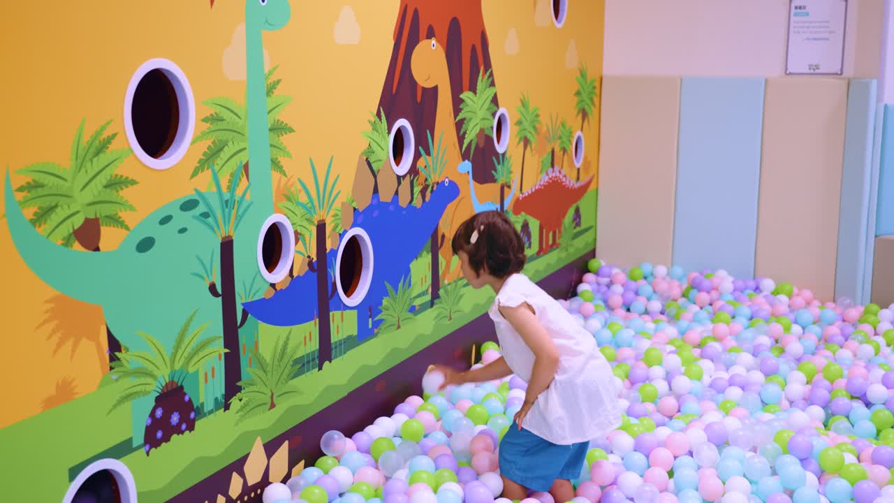 Cute 5 year old mixed Korean girl playing in a colorful ball pit, throwing balls into holes on a dinosaur themed wall - Slow motion
