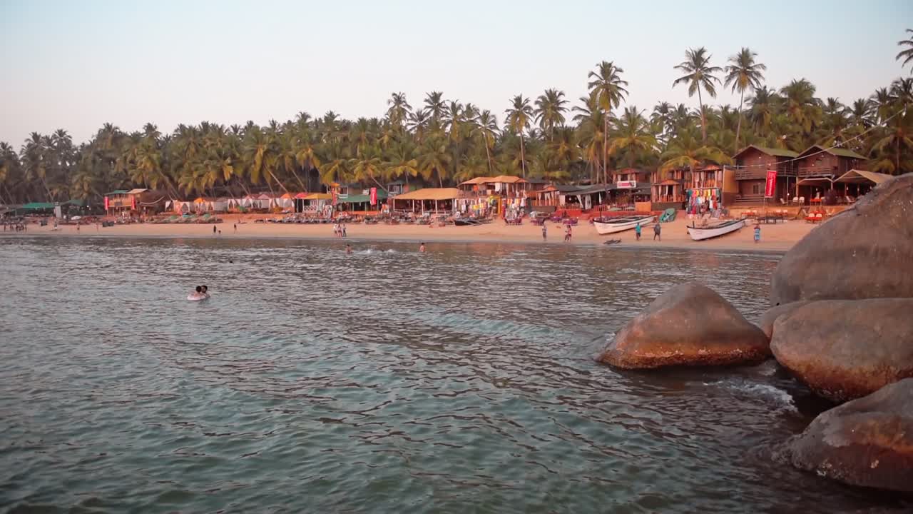 People Enjoying The Cool Sea Water Near The Big Rocks At Palolem Beach In Goa, India With Lush Palm Trees And Boats Docked On The Shore - Wide Shot