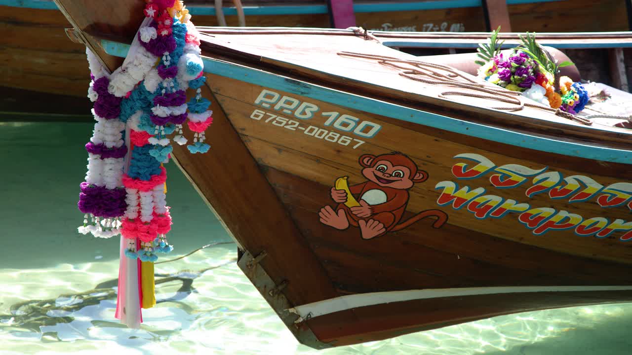 Primer plano de la proa de una barca tradicional de cola larga de madera decorada con guirnaldas vibrantes en agua turquesa.