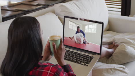 composición de una mujer sentada en casa sosteniendo un café viendo un partido de judo en una computadora portátil