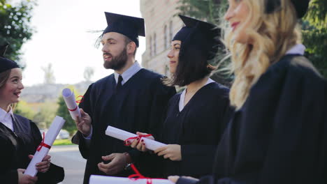 grupo de graduados masculinos y femeninos multiétnicos vestidos con ropa tradicional y gorras con diplomas en las manos hablando y riendo