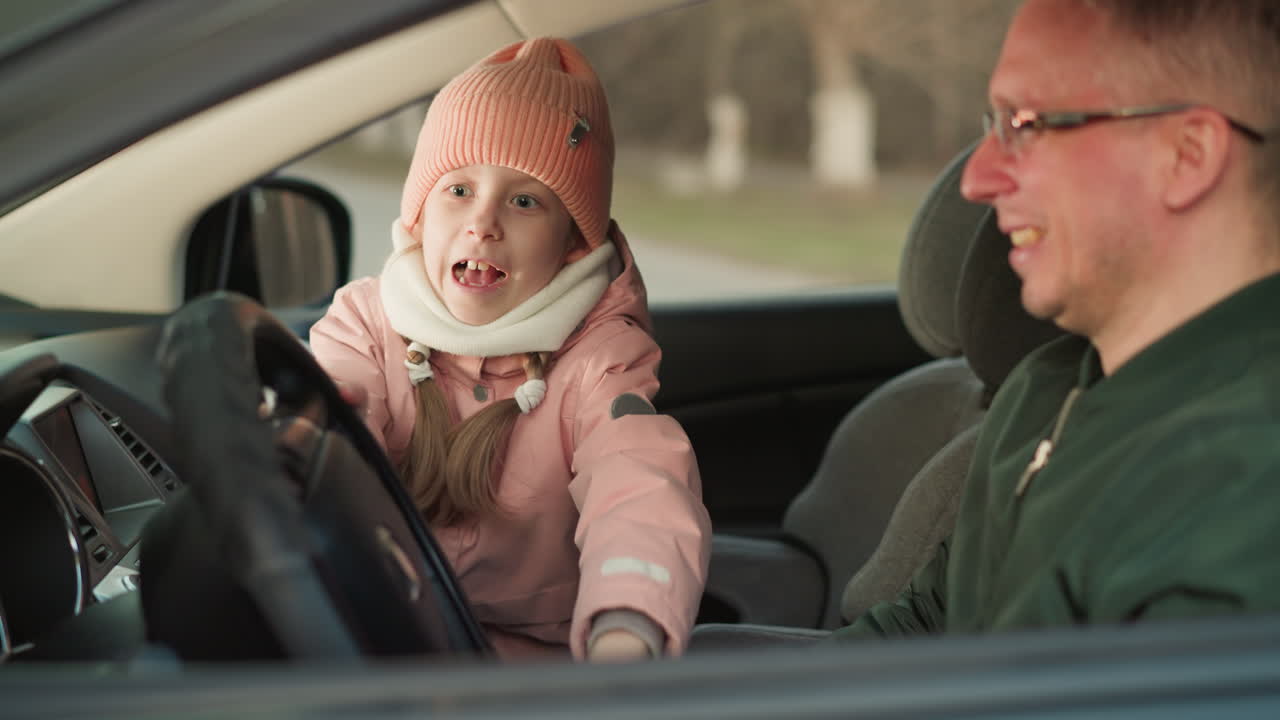 una joven, vestida cálidamente con una gorra rosada y una chaqueta, se sienta en el asiento delantero del pasajero de un coche, girando ansiosamente el volante mientras su padre, sentado a su lado, ríe alegremente