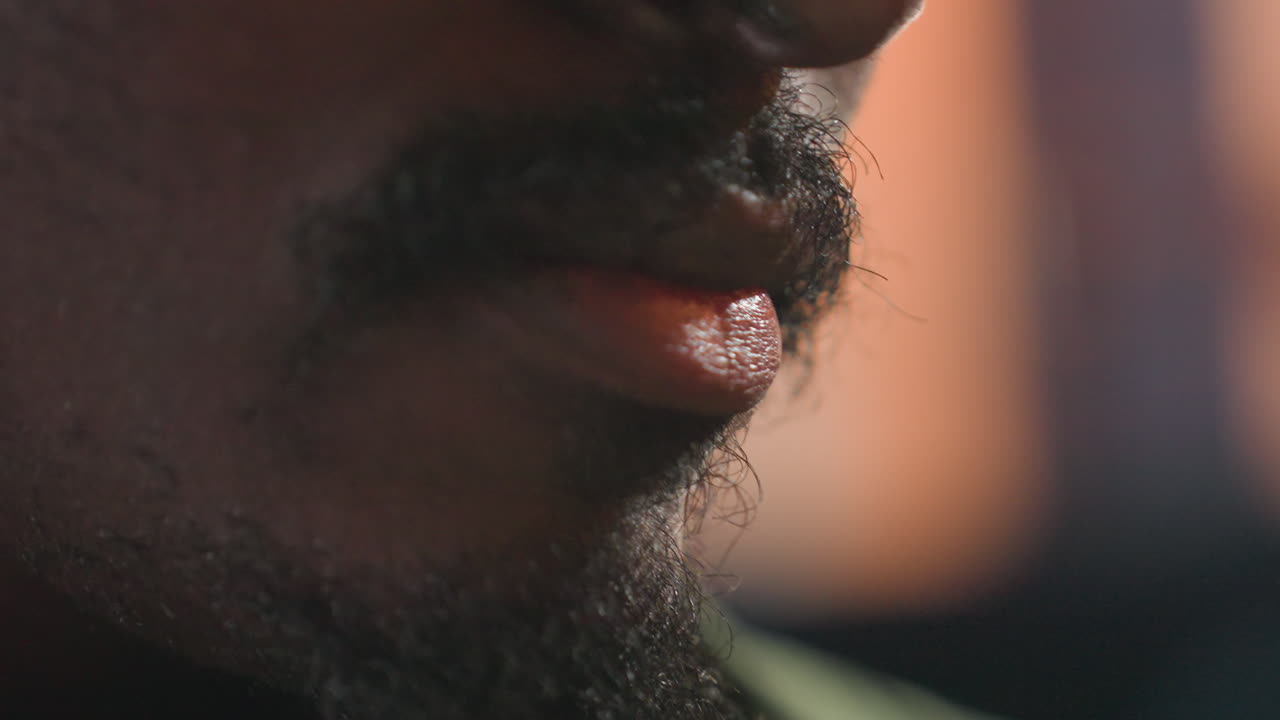 Sortklædt mand smager te i glas, nærbillede af mund og hånd, der rører i teposen, ravfarvet væske og damp, sommelier og barista udforsker aroma og smag, intim caféstemning med varme toner