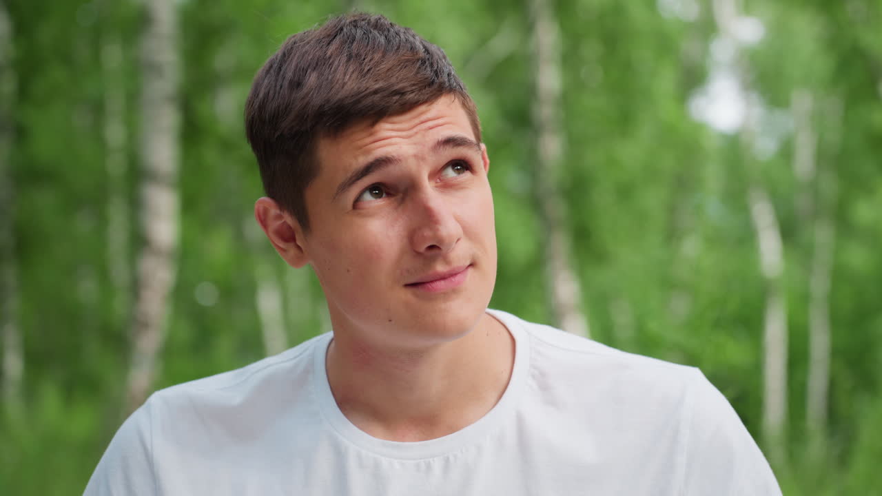 흰 셔츠를 입고 숲에서 무릎을 꿇은 젊은 남자가, 풍부한 녹색 배경과 차분한 자연 조명으로 둘러싸여, 사려 깊은 감정으로 표정 있게 위아래를 바라보는 모습이에요.