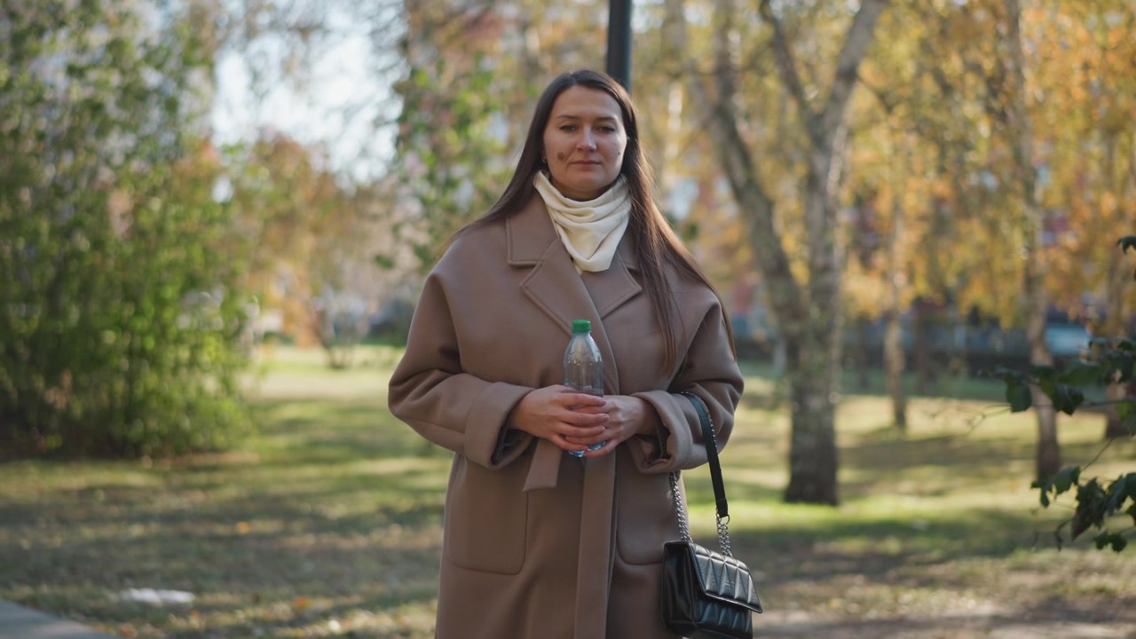 Donna con cappotto beige che tiene una bottiglia d'acqua nel parco