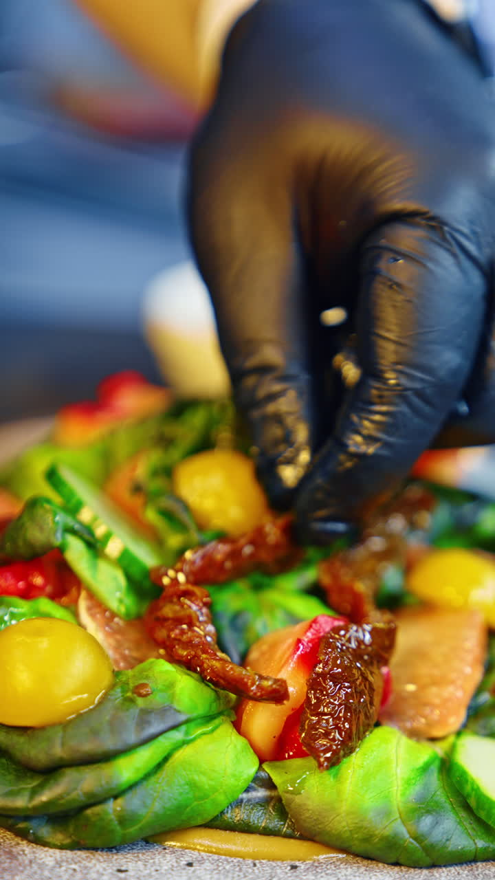Unrecognized cook wearing black latex gloves puts the dried tomatoes on salad leaves. Serving the dish in the restaurant. Close up. Vertical video.