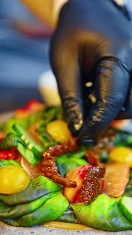 unrecognized cook wearing black latex gloves puts the dried tomatoes on salad leaves. serving the dish in the restaurant. close up. vertical video.
