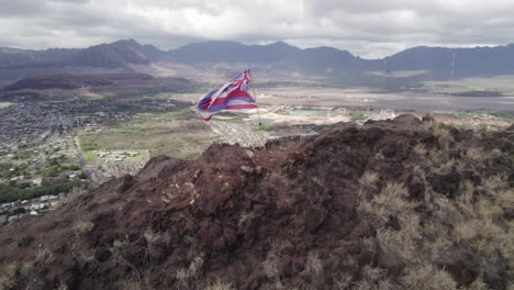 worn hawaiian flag waves on a windswept volcanic ridge, high above a suburban valley. the rugged terrain contrasts with the developed landscape and moody cloud-shrouded mountains beyond