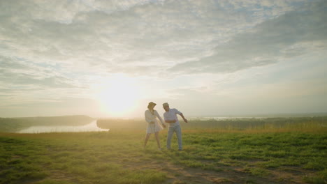 los amantes bailan uno al lado del otro en una colina cubierta de hierba al atardecer cerca de un lago tranquilo. el hombre con una camisa blanca y un sombrero y la mujer con un vestido blanco fluido mueven alegremente sus manos juntos