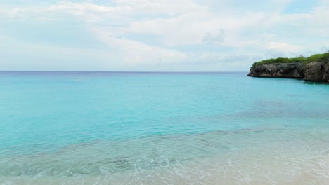tropical beach scene at grote knip in curacao, showcasing clear turquoise waters, palm trees, and pristine sandy beach chairs, aerial dolly over water