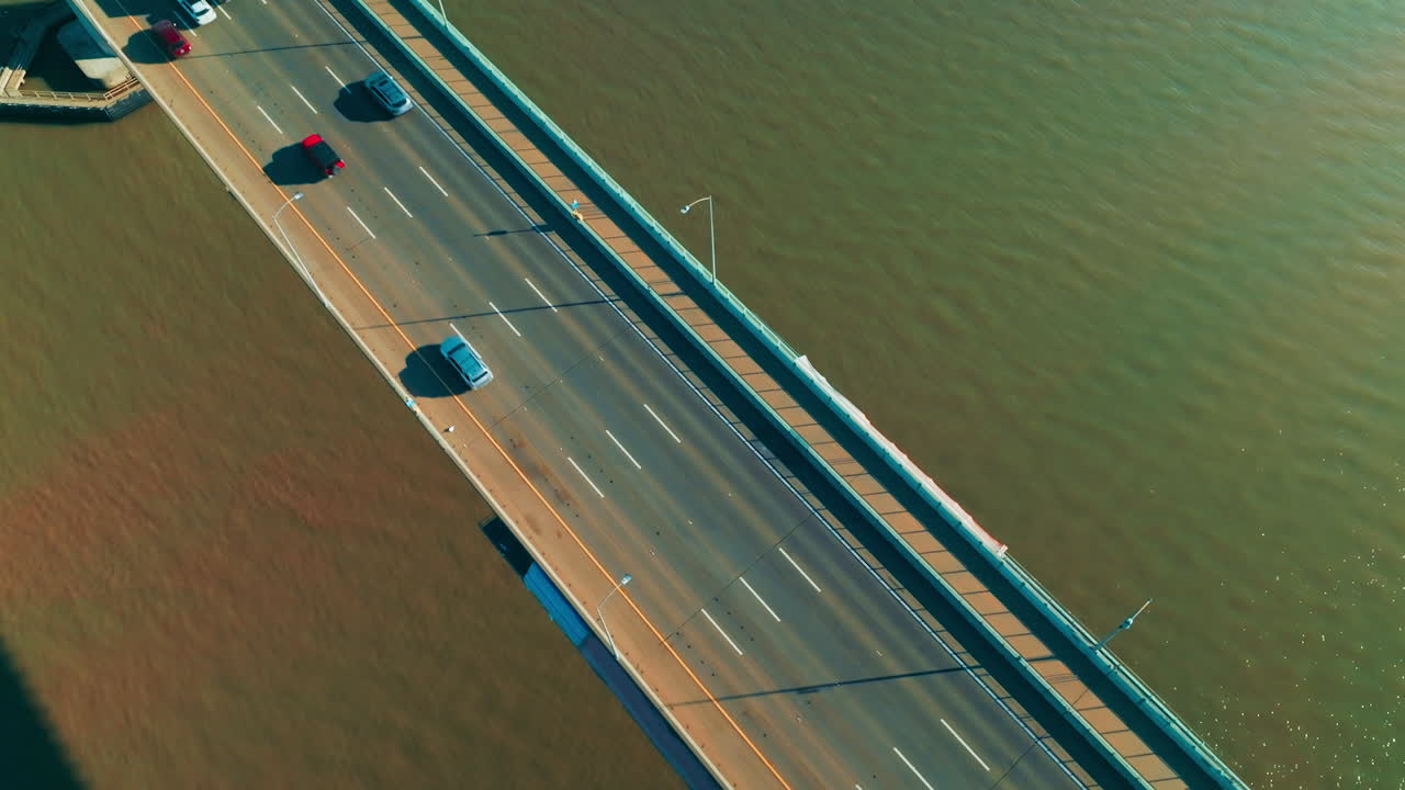Following the cars moving by the bridge with one-way multi-lane highway. Sunny footage over the bridge at backdrop of green water. Top view.