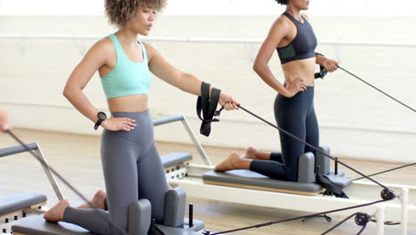 Biracial-woman-using-Pilates-reformer,-Caucasian-woman-adjusting-straps