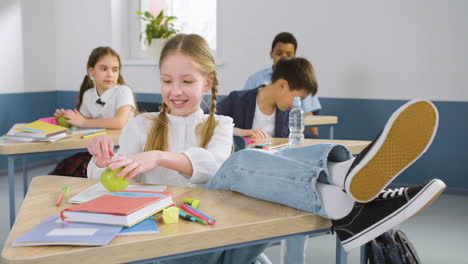 Studentin,-Die-Mit-Ihren-Beinen-Auf-Dem-Tisch-Im-Englischen-Klassenzimmer-Am-Schreibtisch-Sitzt,-Während-Sie-In-Die-Kamera-Schaut-Und-Einen-Apfel-Isst