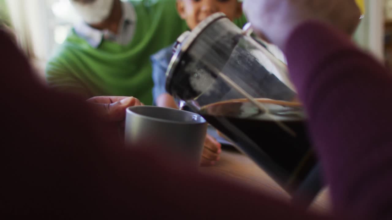 아프리카계 미국인 남자가 집에서 가족과 함께 아침 식사를 하는 동안 컵에 커피를  ⁇ 고 있습니다.