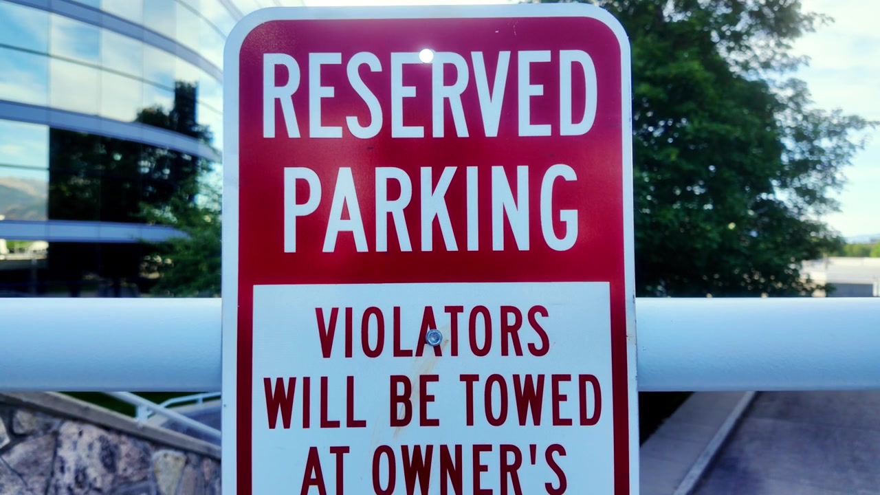 Cartello rosso e bianco con la scritta "Parcheggio riservato. I trasgressori saranno rimossi a spese del proprietario", con edifici di un complesso commerciale sullo sfondo, ripresa frontale diretta in un parcheggio con alberi sullo sfondo.