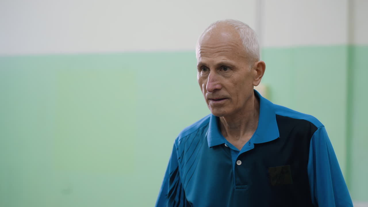 Close up of senior man in blue shirt actively playing table tennis while interacting, showing focus, energy, and communication during dynamic indoor training session with paddle and ball