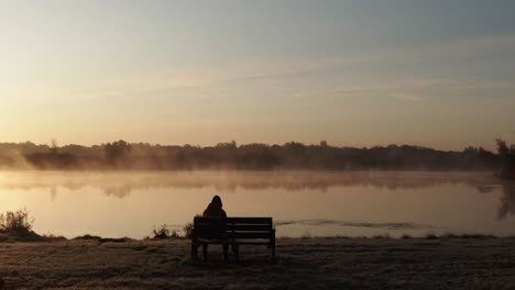 Vista-Trasera-De-Una-Persona-Solitaria-Sentada-En-Un-Banco-Frente-A-Un-Lago-Brumoso-Durante-La-Puesta-De-Sol,-Concepto-Triste-Y-Deprimente